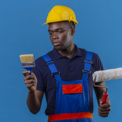 young african american builder man wearing construction uniform and safety helmet holding brush and paint roller looking at brush with serious expression standing over blue background young african american builder man wearing construction uniform and safety helmet holding brush and paint roller looking at brush with serious expression standing over blue background